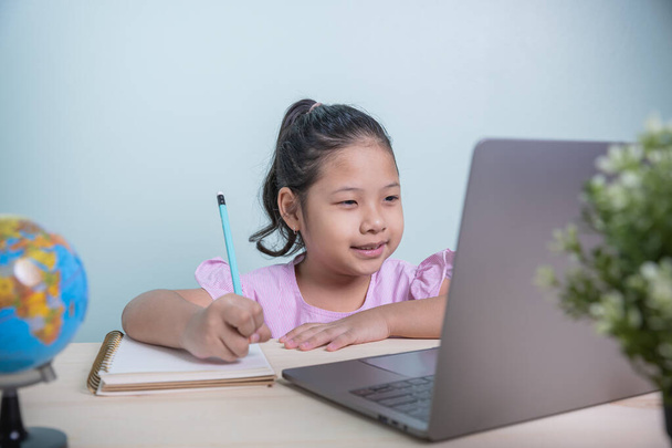 アジアの小さな子供の女の子は、ビデオでオンラインレッスンを学ぶ部屋のラップトップからインターネットを呼び出します。コロナウイルスの普及により、学生は自宅からオンラインで勉強することを余儀なくされました. - 写真・画像