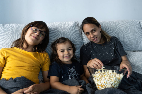 Mãe e filhas caucasianas, sentadas no sofá da sala, assistindo a um filme enquanto comem pipocas, - Foto, Imagem