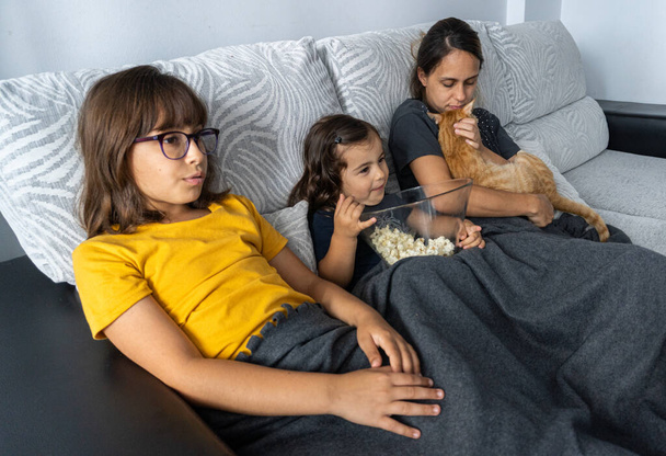 Caucasian mother and daughters, sitting on the living room couch, watching a movie while eating popcorn, - Photo, Image