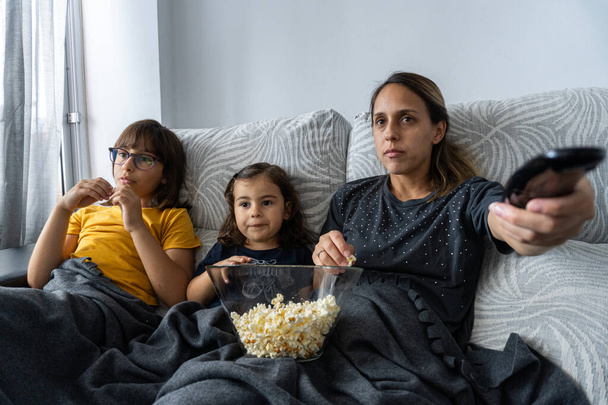 Mãe e filhas caucasianas, sentadas no sofá da sala, assistindo a um filme enquanto comem pipocas, - Foto, Imagem