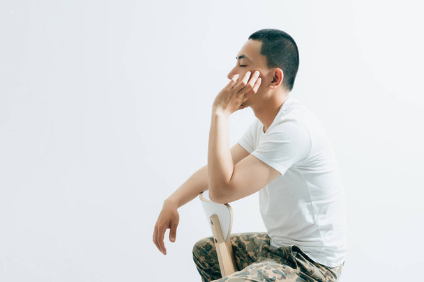 Schöner asiatischer Typ in Camouflage-Hose und weißem T-Shirt auf einem Stuhl sitzend. - Foto, Bild
