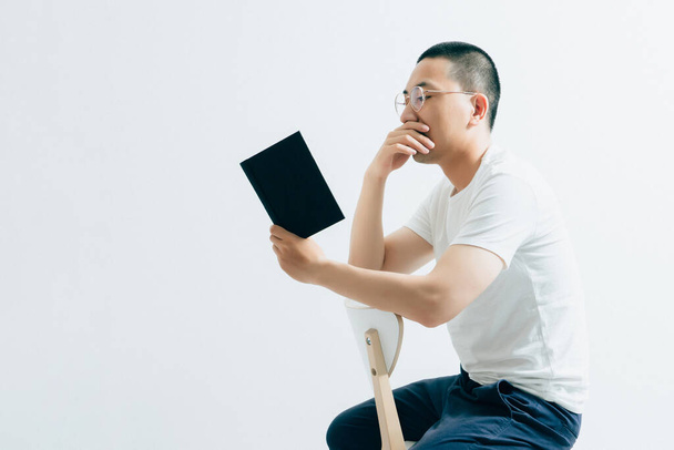 Un guapo asiático leyendo un libro sentado frente a un fondo blanco. - Foto, imagen