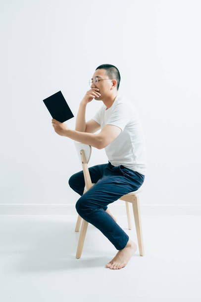 Un bel ragazzo asiatico che legge un libro seduto davanti a uno sfondo bianco. - Foto, immagini