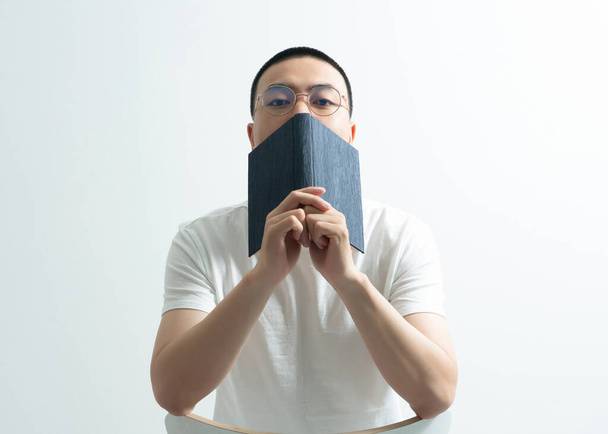 Ein hübscher asiatischer Typ liest ein Buch und sitzt vor weißem Hintergrund. - Foto, Bild