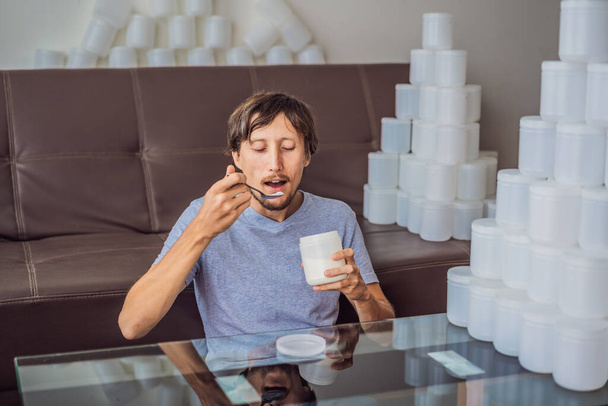 The man eats yogurt and is surrounded by many jars of yoghurt that he uses in a year. A huge amount of plastic food jars. Excessive consumption of plastic. Reduce, reuse, recycle - Photo, Image