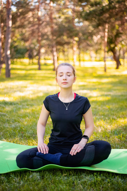 A young woman dressed in a sports uniform sits in a lotus position on a fitness mat in a city park. - Foto, afbeelding