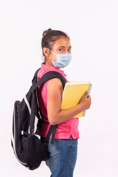 Kis latin lány egy arc maszk visel egy nagy hátizsák néz vissza a kamerába: iskolai koncepció. - Fotó, kép