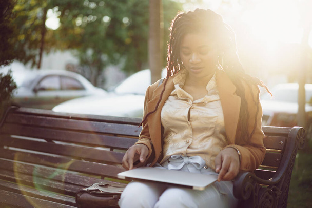 African american woman freelancer sitting on a bench in a street and using laptop - 写真・画像