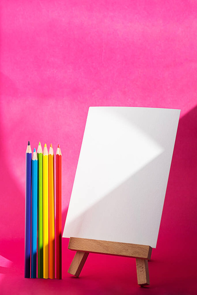 ミニイーゼルと色鉛筆上のブランクシートペーパー。空の白いページ創造的な概念は学校に戻る。文房具まだ人生上の明るいピンクの背景. - 写真・画像