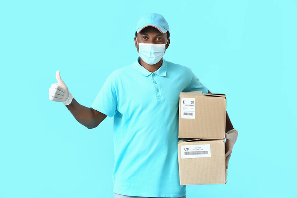 Afro-Amerikaanse bezorger in medisch masker en met pakketten met duim-up op kleur achtergrond - Foto, afbeelding