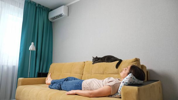 Жінка вмикає кондиціонер за допомогою пульта дистанційного керування, лежить на дивані з кішкою
 - Фото, зображення