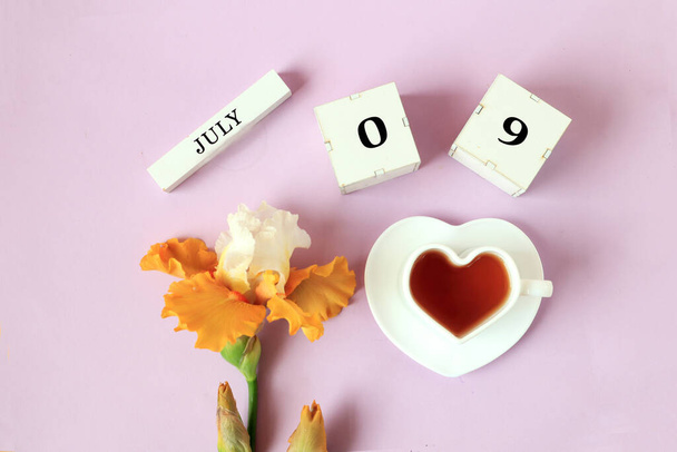 Calendar for July 9 : the name of the month of July in English, cubes with the numbers 0 and 9, a cup of tea in the shape of a heart, yellow iris on a pastel background, top view - Φωτογραφία, εικόνα