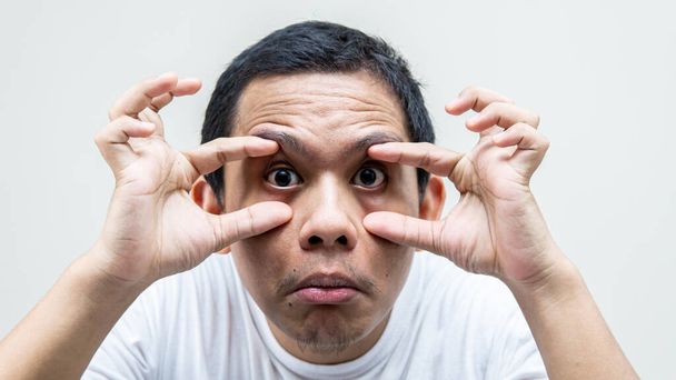 Een portret van een jonge Maleise man die gedwongen werd zijn ogen te openen met zijn vinger op geïsoleerde witte achtergronden. Ik voel me moe, slaperig.. - Foto, afbeelding