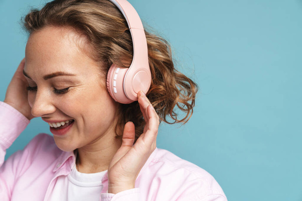 Young ginger woman laughing while listening music with headphones isolated over blue background - Foto, afbeelding