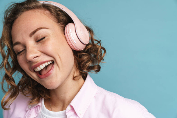Young ginger woman laughing while listening music with headphones isolated over blue background - Foto, afbeelding