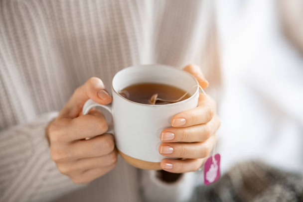 Woman wearing sweater and holding a warm cup of freshly brewed tea. Close up of woman hands holding mug of hot detox infusion in morning. Girl with cozy cloths relaxing at home during tea time in cold winter day. - Foto, imagen