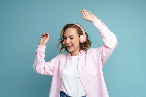 Young ginger woman dancing while listening music with headphones isolated over blue background - Foto, afbeelding