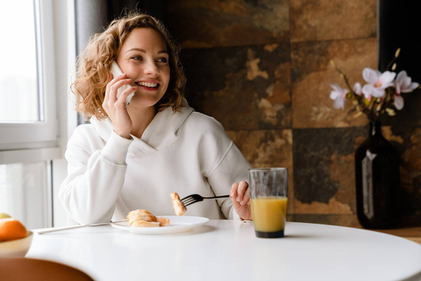 Young white woman talking on mobile phone while having breakfast at home - Valokuva, kuva