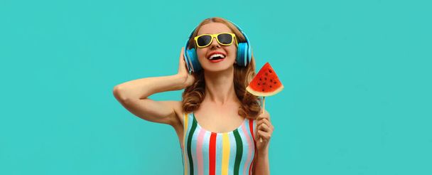Summer colorful portrait of cheerful happy laughing young woman in headphones listening to music with juicy lollipop or ice cream shaped slice of watermelon on blue background - Φωτογραφία, εικόνα