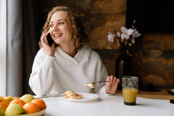Молодая белая женщина разговаривает по мобильному телефону за завтраком дома - Фото, изображение