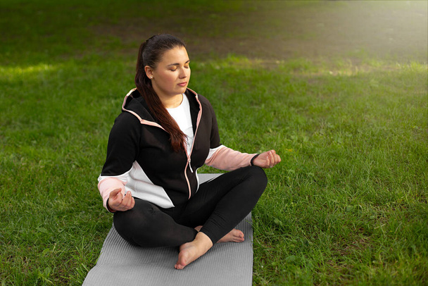 beautiful girl meditates in the lotus position on the green grass surrounded by trees. the yoga class in nature. mental health concept - Photo, Image