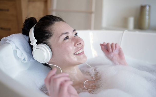Happy smiling young multicultural asian woman with headphones enjoying a hot bath. Multi ethnic millennial girl listening music relaxing in bathroom, treatment therapy healthy lifestyle concept - Фото, изображение