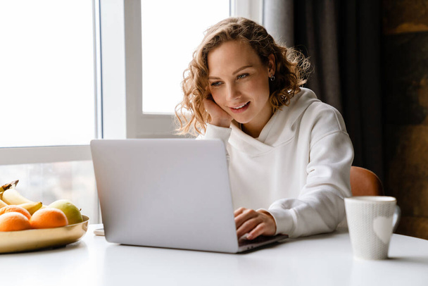 White blonde woman smiling and typing on laptop while sitting at table in home - Valokuva, kuva
