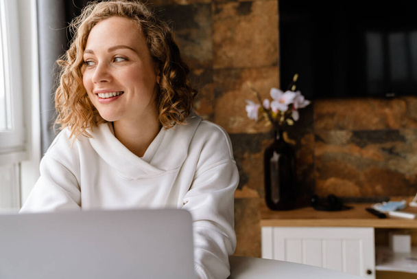 White blonde woman smiling and typing on laptop while sitting at table in home - Photo, Image