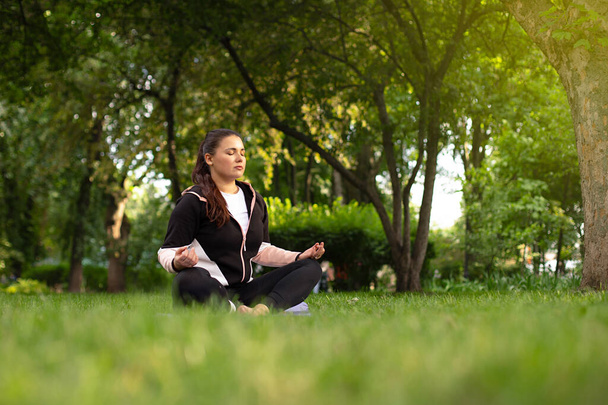 menina bonita medita na posição de lótus na grama verde cercada por árvores. a aula de ioga na natureza. conceito de saúde mental - Foto, Imagem
