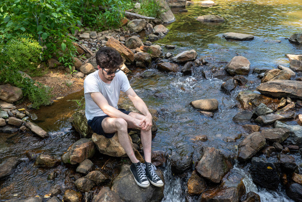 Smutny młody człowiek siedzi na stopniach w strumieniu płynącym patrząc w dół - Zdjęcie, obraz