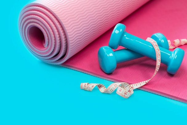 En una esterilla de yoga rosa hay pesas azules y una cinta métrica. Composición brillante sobre un fondo azul, mucho espacio libre para su diseño. Dieta, pérdida de peso y el concepto de acondicionamiento físico para las mujeres. - Foto, Imagen
