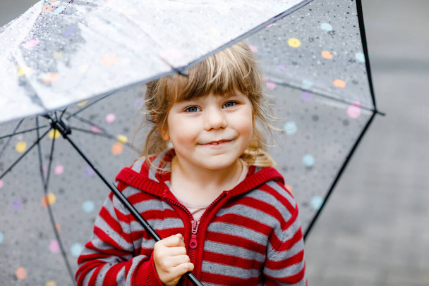 Little toddler girl playing with big umbrella on rainy day. Happy positive child running through rain, puddles. Preschool kid with rain clothes and rubber boots. Children activity on bad weather day. - Photo, Image