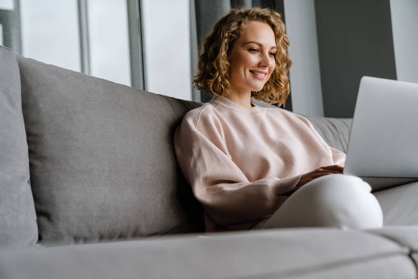 White blonde woman smiling and using laptop while sitting on couch at home - Foto, imagen