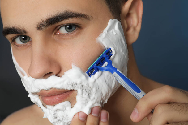 Handsome young man shaving with razor on blue background, closeup - Foto, Imagen