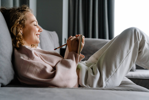 Young white woman smiling and writing down notes while lying on couch at home - Photo, image