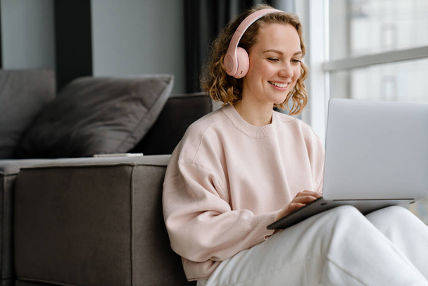 Fehér nő fülhallgatóban laptoppal, miközben otthon ül a padlón - Fotó, kép