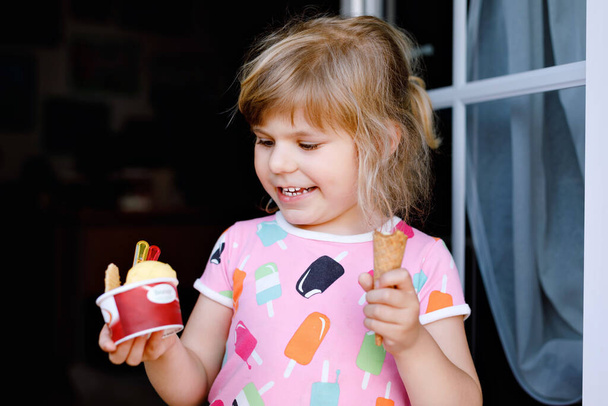 Cute funny preschool girl eating fruit ice cream at home. Happy healthy toddler child smiling and tasting ice-cream in waffle cone. Lovely kid enjoying summer dessert. - Photo, Image