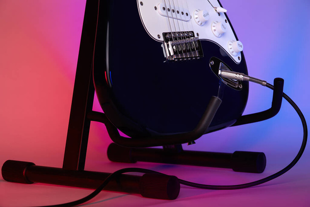 Uma guitarra elétrica em um suporte conectado com um cabo a um amplificador é iluminada com luz azul e vermelha. Close-up. - Foto, Imagem