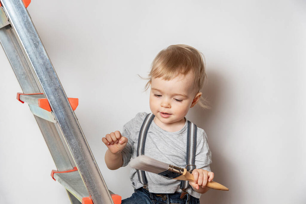 Маленький милый красивый ребенок рядом с лестницей с кистью в руках на фоне белой стены.  - Фото, изображение