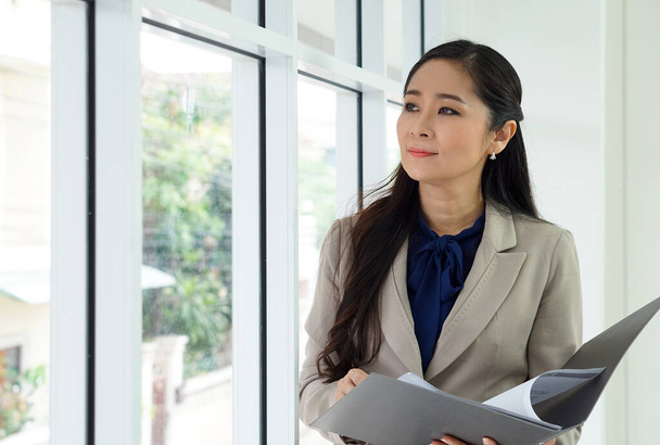 Ázsiai női irodavezető barna öltönyös irattartóval a kezében. Kinézett, miközben gondolt valamire.. - Fotó, kép