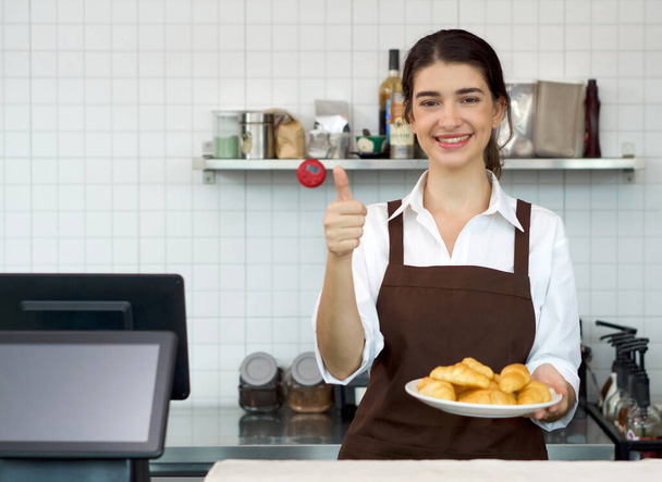 Nuori kaukasialainen kauppias hymyilee ja nostaa peukalot ylös pitäen croissantin lautasta kahvilan tiskin edessä. Aamuilmapiiri kahvilassa. - Valokuva, kuva