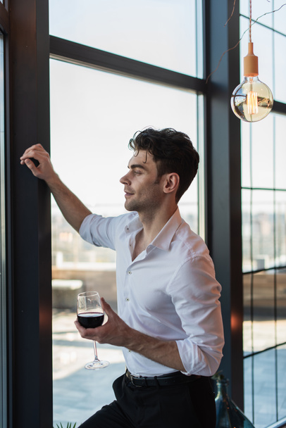 elegant man in white shirt holding glass of red wine while standing near window - Photo, Image