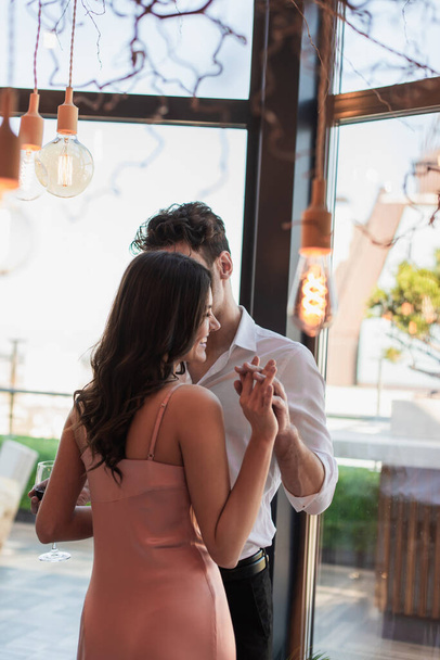 man and cheerful woman holding hands in restaurant near decorative light bulbs  - 写真・画像