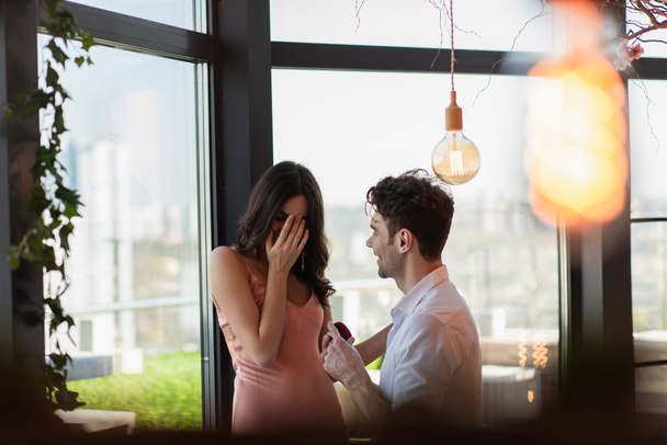 happy man making proposal to girlfriend in slip dress covering face in restaurant  - Фото, изображение