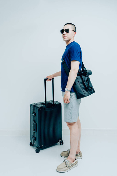 Koyu mavi tişörtlü ve şortlu genç bir adam bavulu çekiyor. - Fotoğraf, Görsel