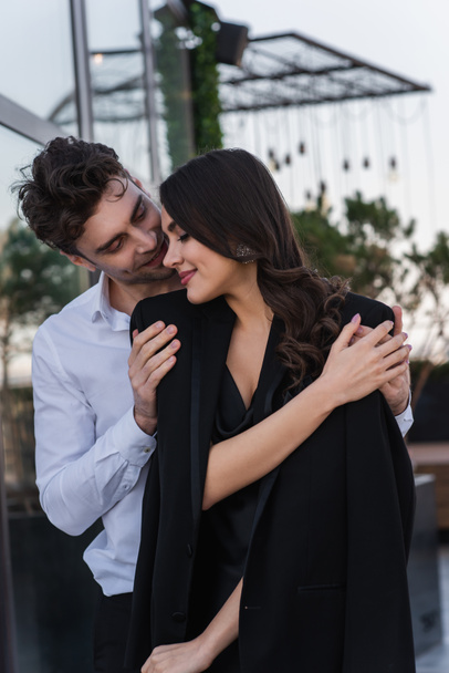 elegante man knuffelen vrolijke vrouw in zwart blazer  - Foto, afbeelding