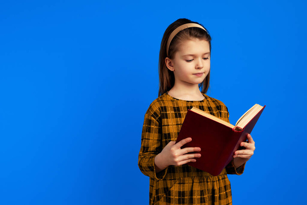Serious little girl reading book while standing against blue background  - Фото, изображение