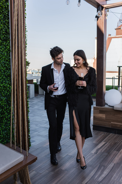full length of elegant woman and man smiling while holding glasses of wine on terrace  - Фото, изображение