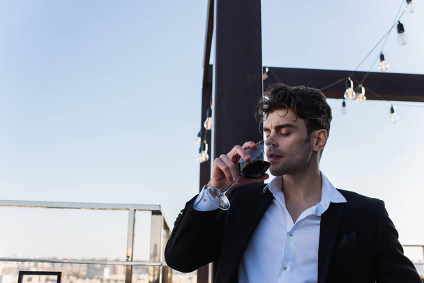 κομψό άνθρωπος με κοστούμι κρατώντας γυαλί και πίνοντας κόκκινο κρασί στην οροφή  - Φωτογραφία, εικόνα