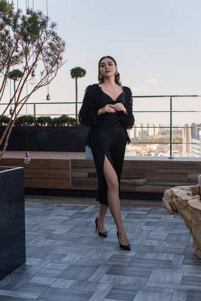 full length of elegant young woman in black slip dress and faux fur jacket walking on roof - Photo, Image